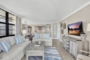 A spacious living room with a large window, a sofa set, a coffee table, and a TV on a wooden cabinet. - Beachfront Condo | Pool, Gym & Tennis Courts (Marco Island)