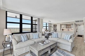 A spacious living room with a large window, a sofa set, a coffee table, and a kitchen area in the background.