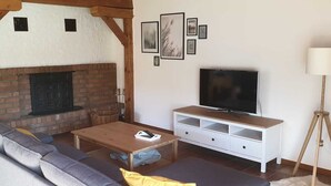 Ein Wohnzimmer mit Kamin aus Backstein, einem großen Sofa, einem Couchtisch, einem Fernseher auf einem weißen Schrank und einer Lampe.