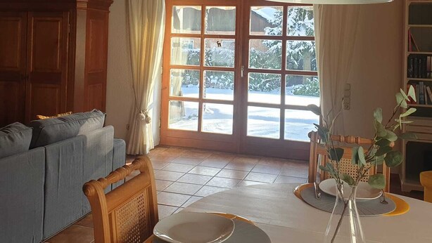 Ein gemütliches Wohnzimmer mit Holzbalkendecke, einem großen Fenster mit Blick auf eine verschneite Landschaft, einer Couch, einem Esstisch mit Tellern und einem Bücherregal.