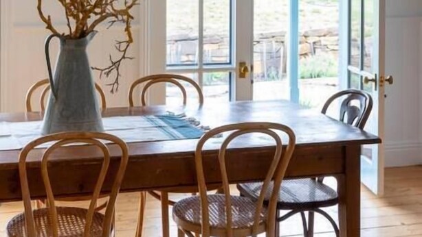 A dining area with a wooden table and wicker chairs, a vase with branches, and a white pendant light.