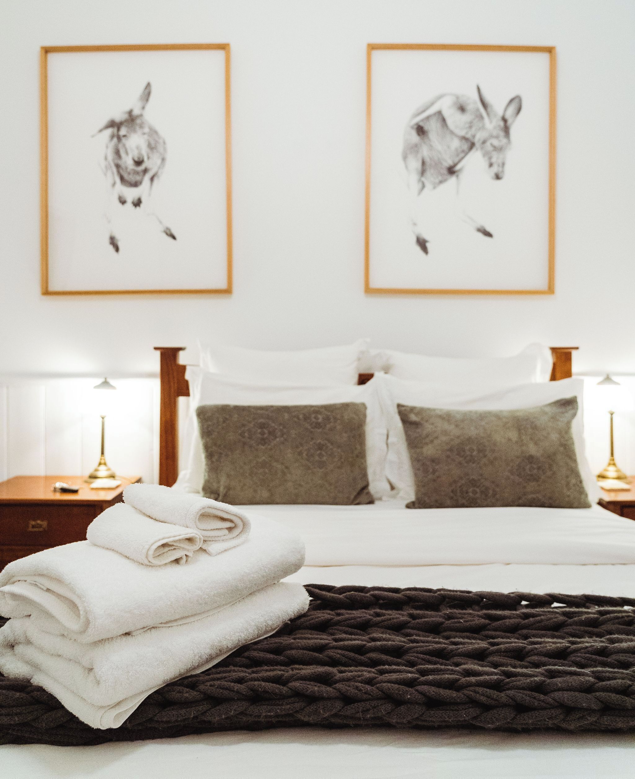 A neatly made bed with white linens, a dark brown throw blanket, and two framed animal prints on the wall.