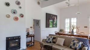 A cozy living room with a fireplace, a sofa with patterned cushions, a dining table with chairs, and a large window.