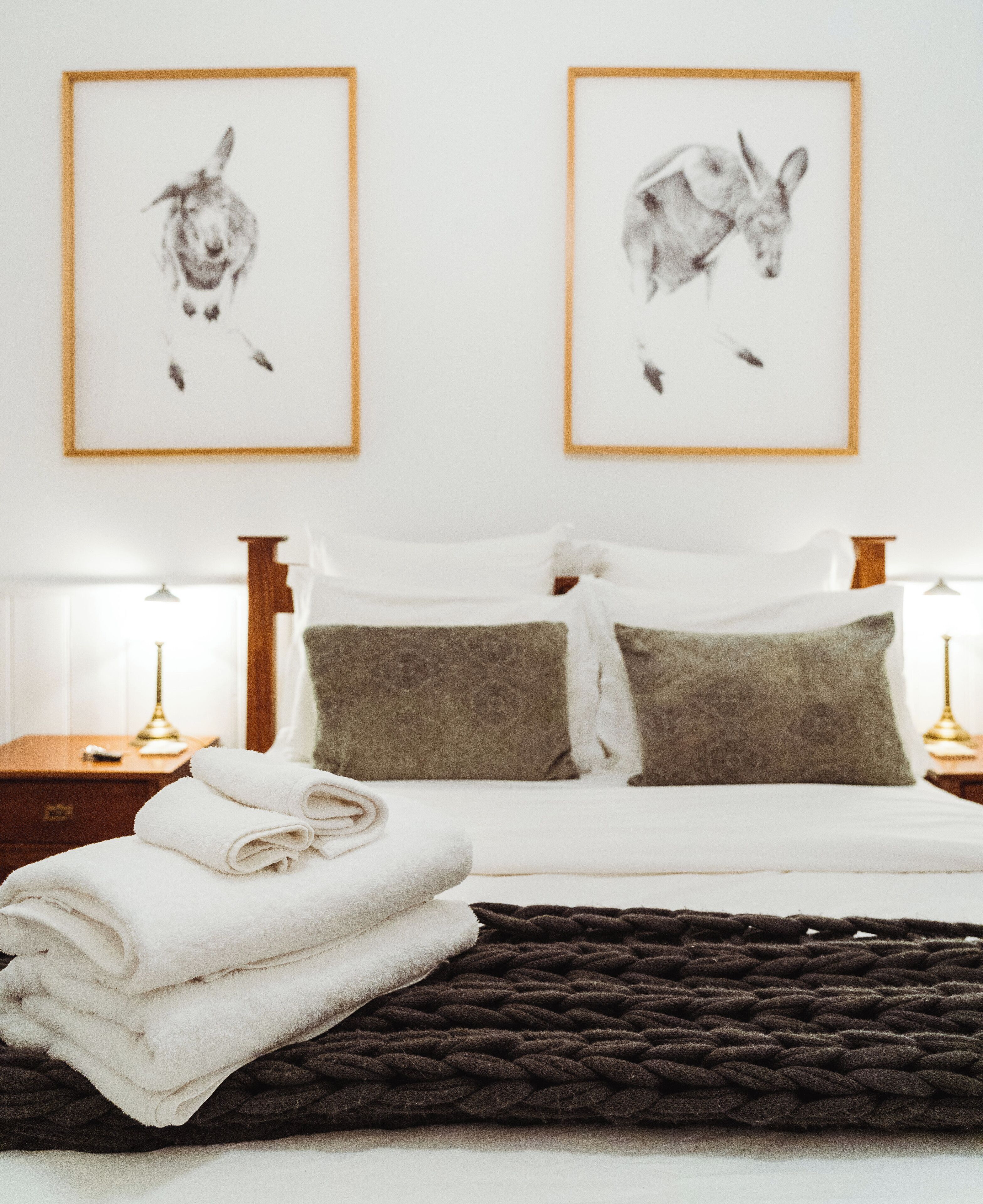 Una cama bien tendida con sábanas blancas, un plaid de punto marrón oscuro y dos cuadros con estampados de animales en la pared.