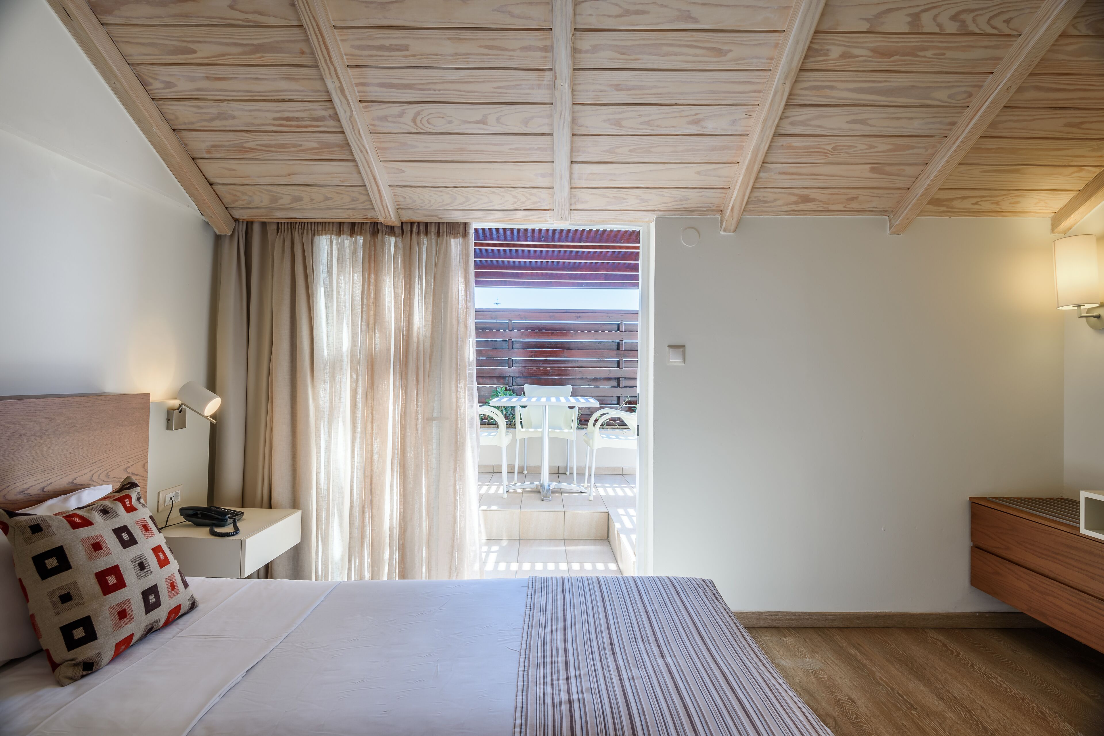 A hotel room with a wooden ceiling, a bed with a patterned pillow, a bedside table with a lamp, and a view of an outdoor patio with a table and chairs.