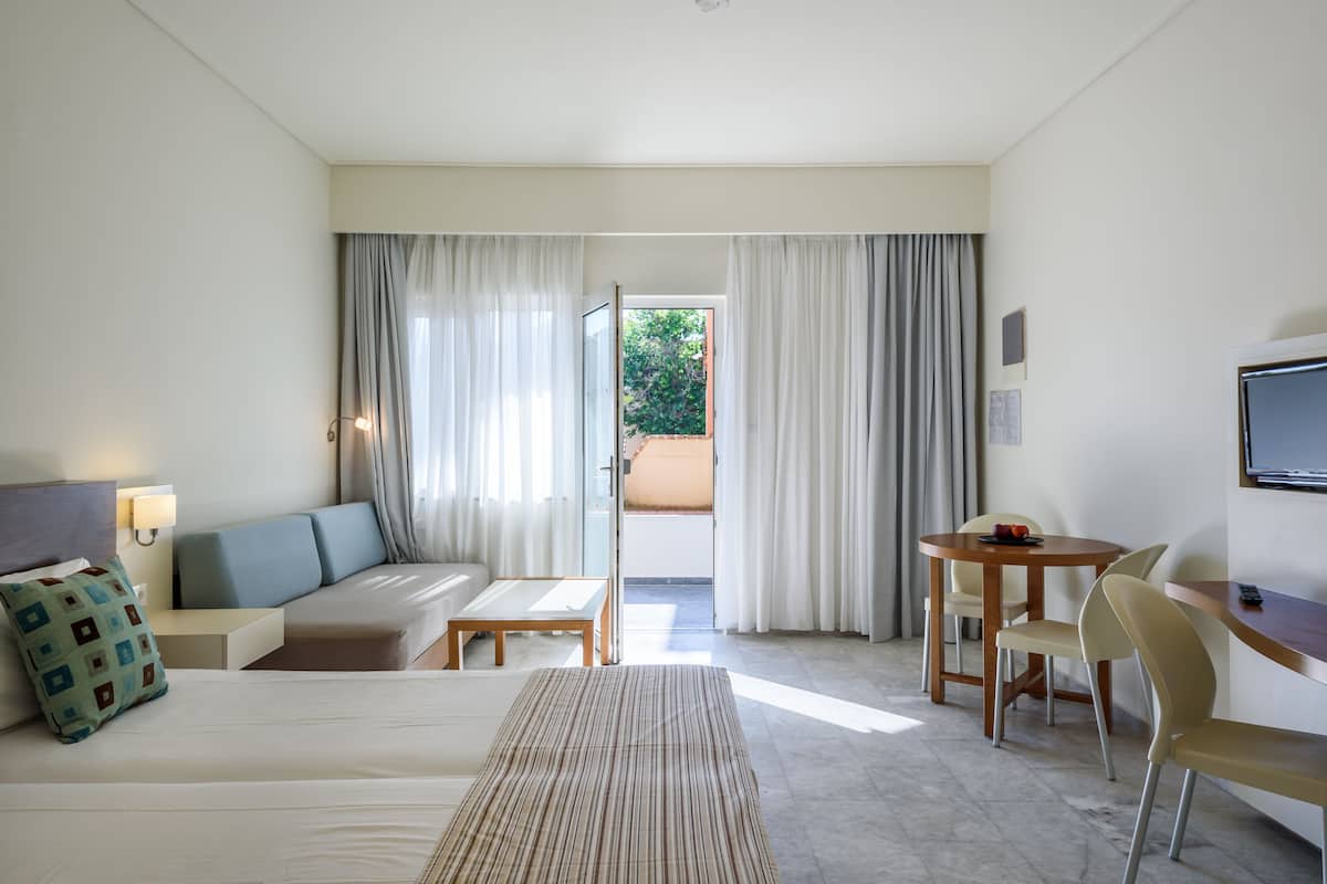 A hotel room with a bed, a sofa, a small table, a chair, a TV, and a balcony with curtains.