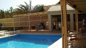 A swimming pool with a wooden deck and chairs, a covered seating area, and a bar.