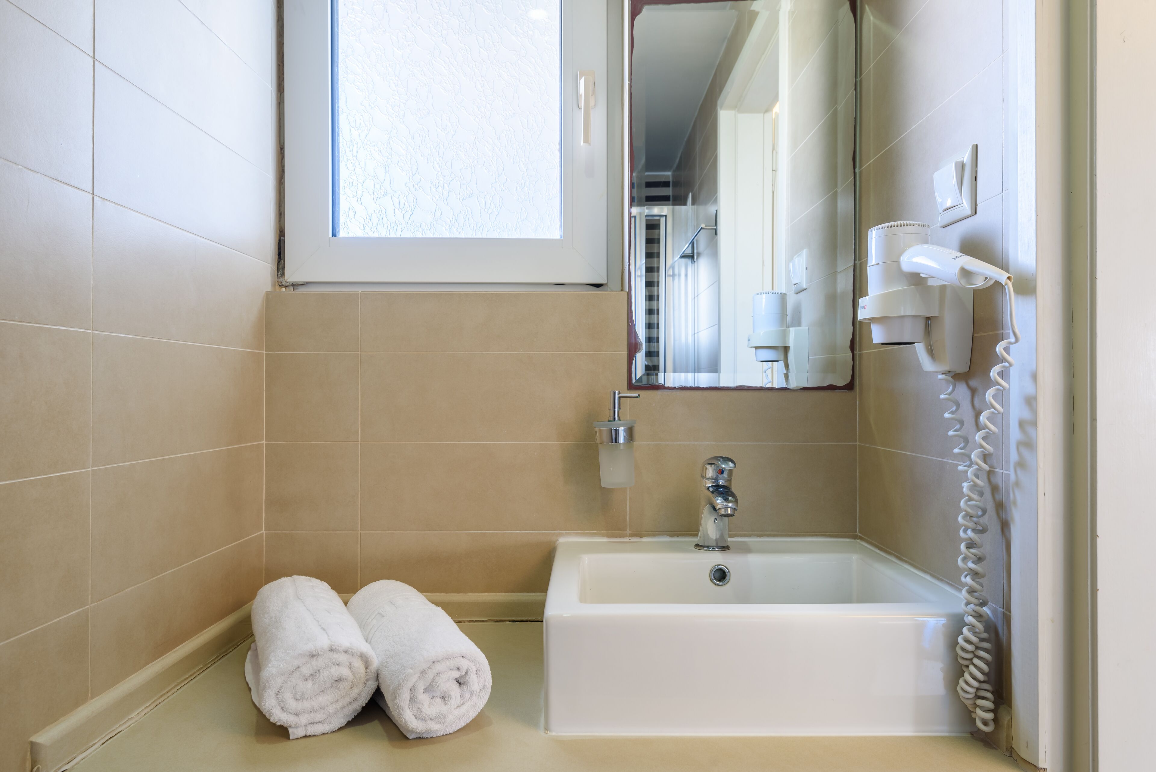 A bathroom with a white sink, a towel rack with two towels, a hairdryer, and a window with blinds.