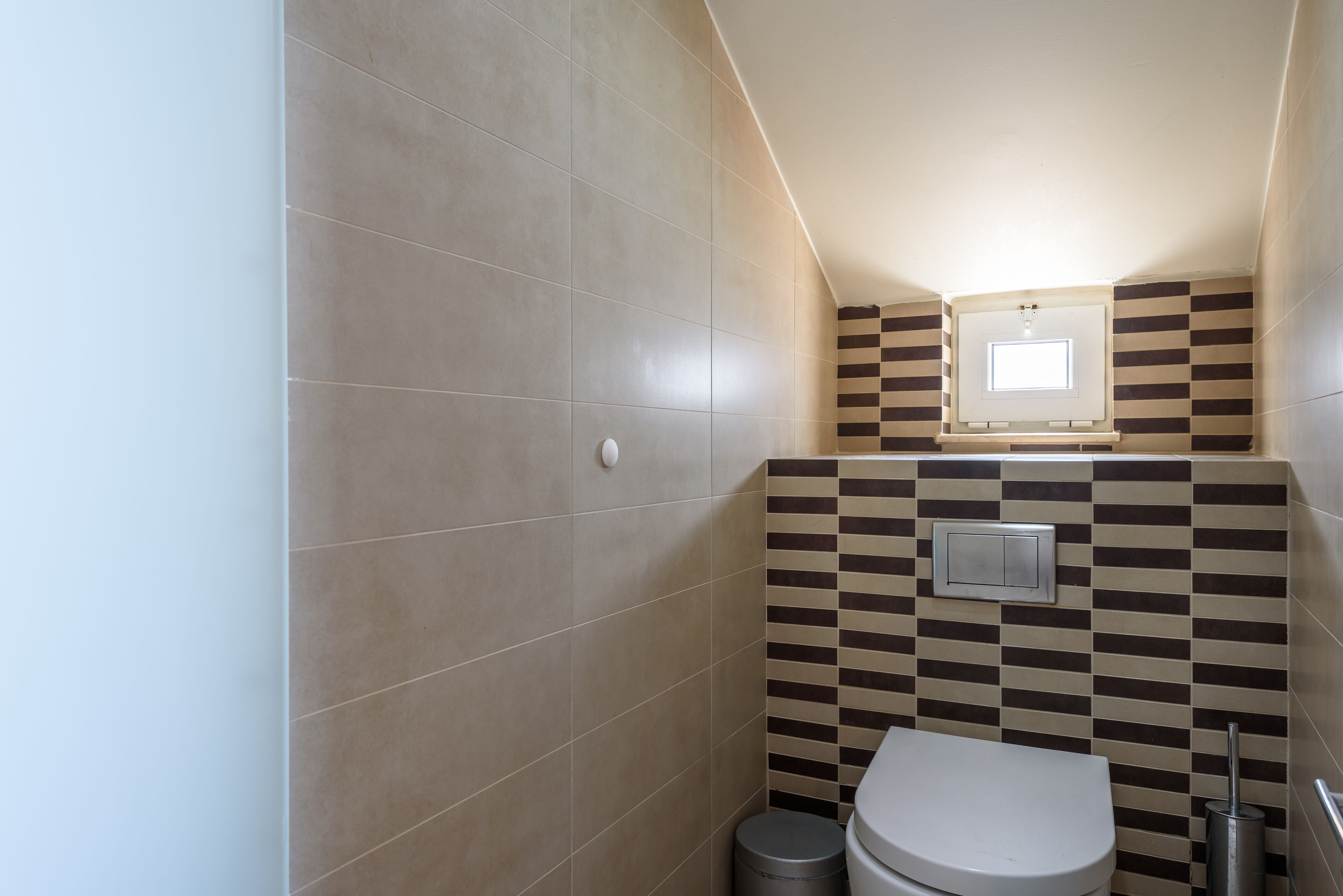 A small, modern bathroom with a toilet, a trash bin, and a wall-mounted light fixture.
