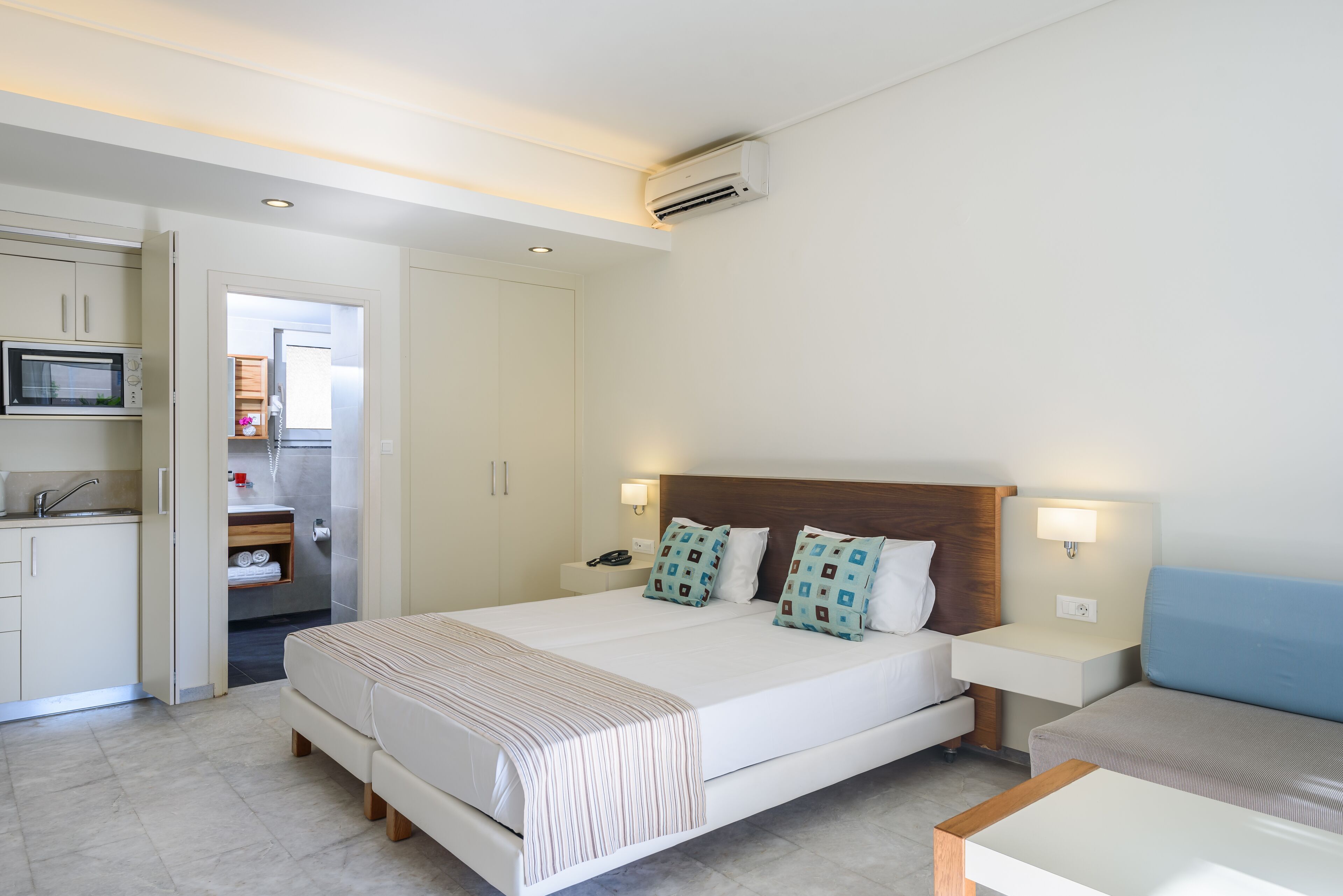 A modern hotel room with a large bed, bedside tables, a built-in TV, and a kitchenette.