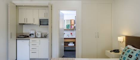 A hotel room with a kitchenette, a bed with a striped bedspread, a nightstand with a lamp, and a view into the bathroom.