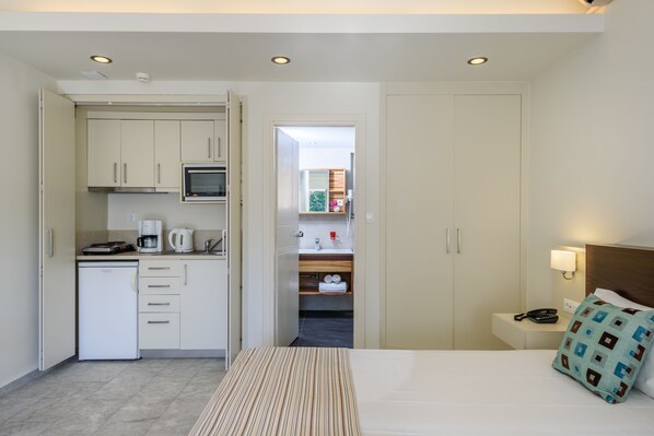 A hotel room with a kitchenette, a bed with a striped bedspread, a nightstand with a lamp, and a view into the bathroom.