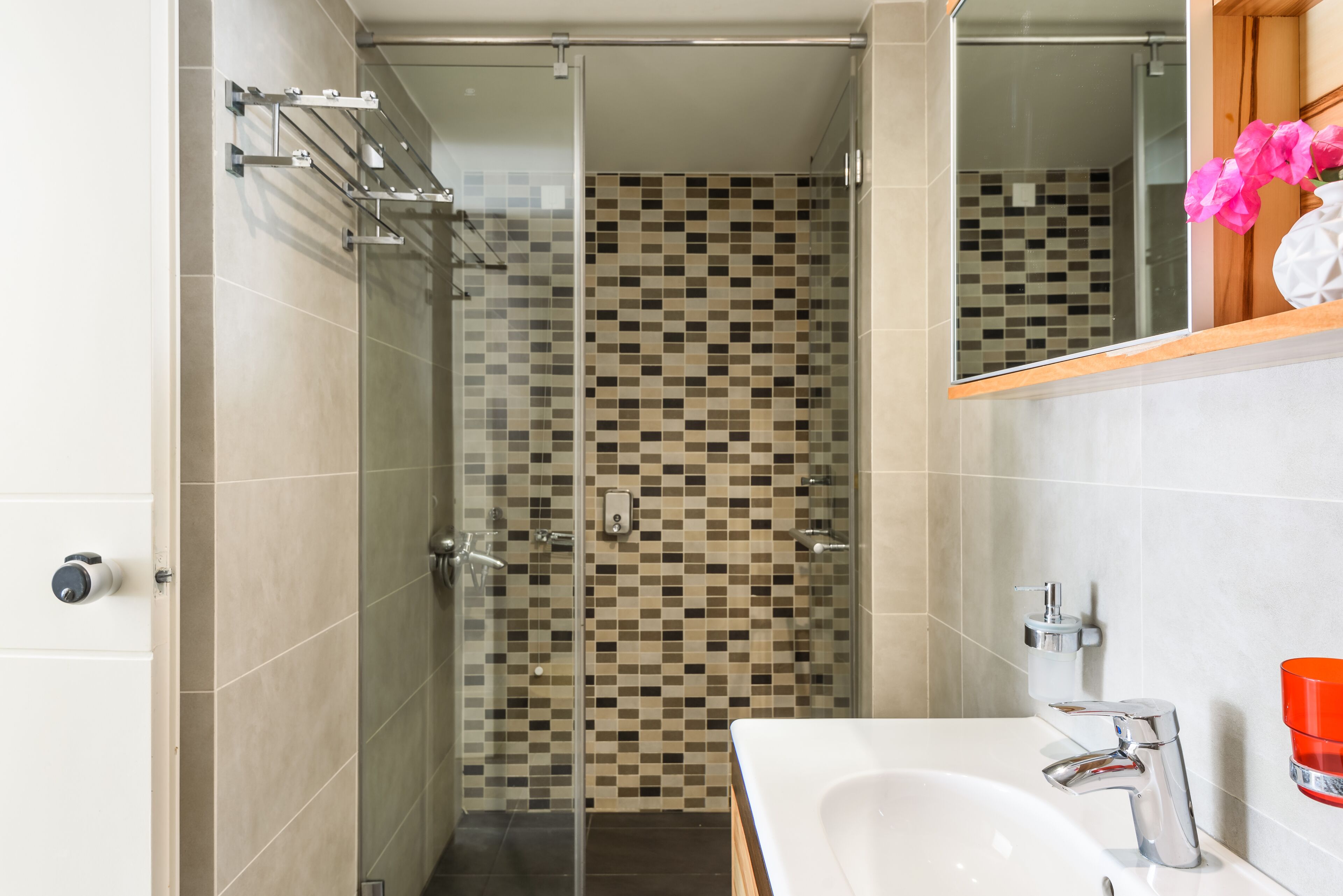 A modern bathroom with a glass shower enclosure, a tiled mosaic backsplash, a white sink with a chrome faucet, and a wooden vanity.