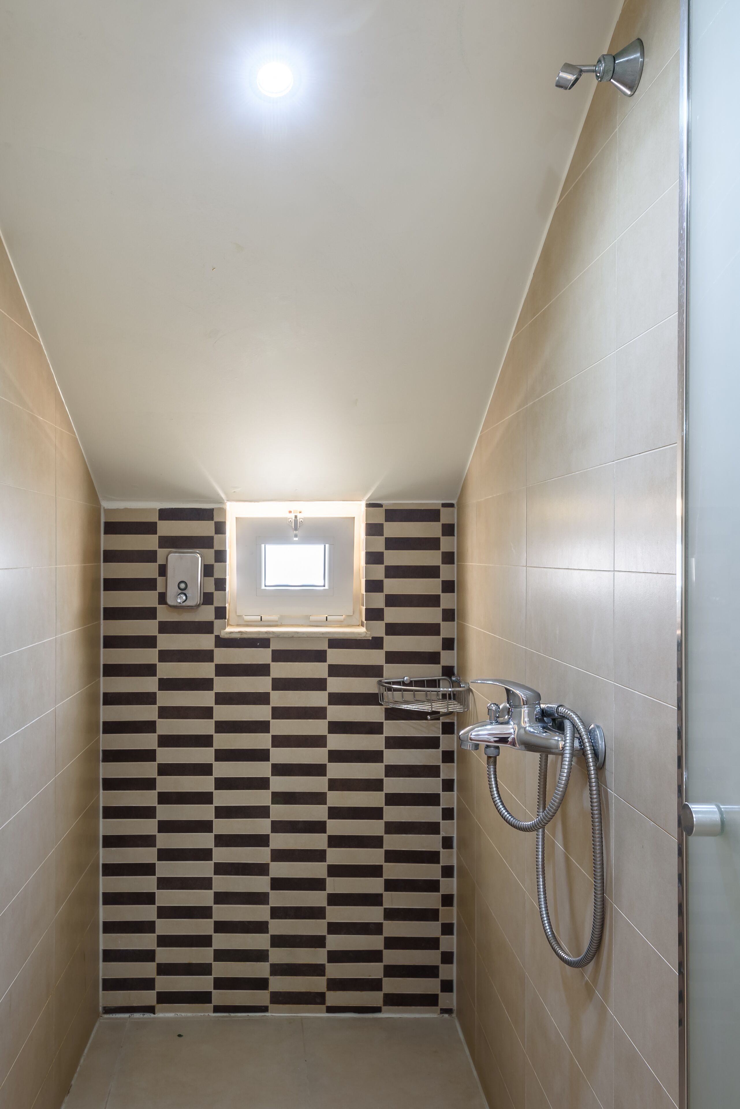 Une petite salle de bain carrelée, avec un motif à carreaux sur un des murs, une fenêtre et une douche équipée d’un flexible.