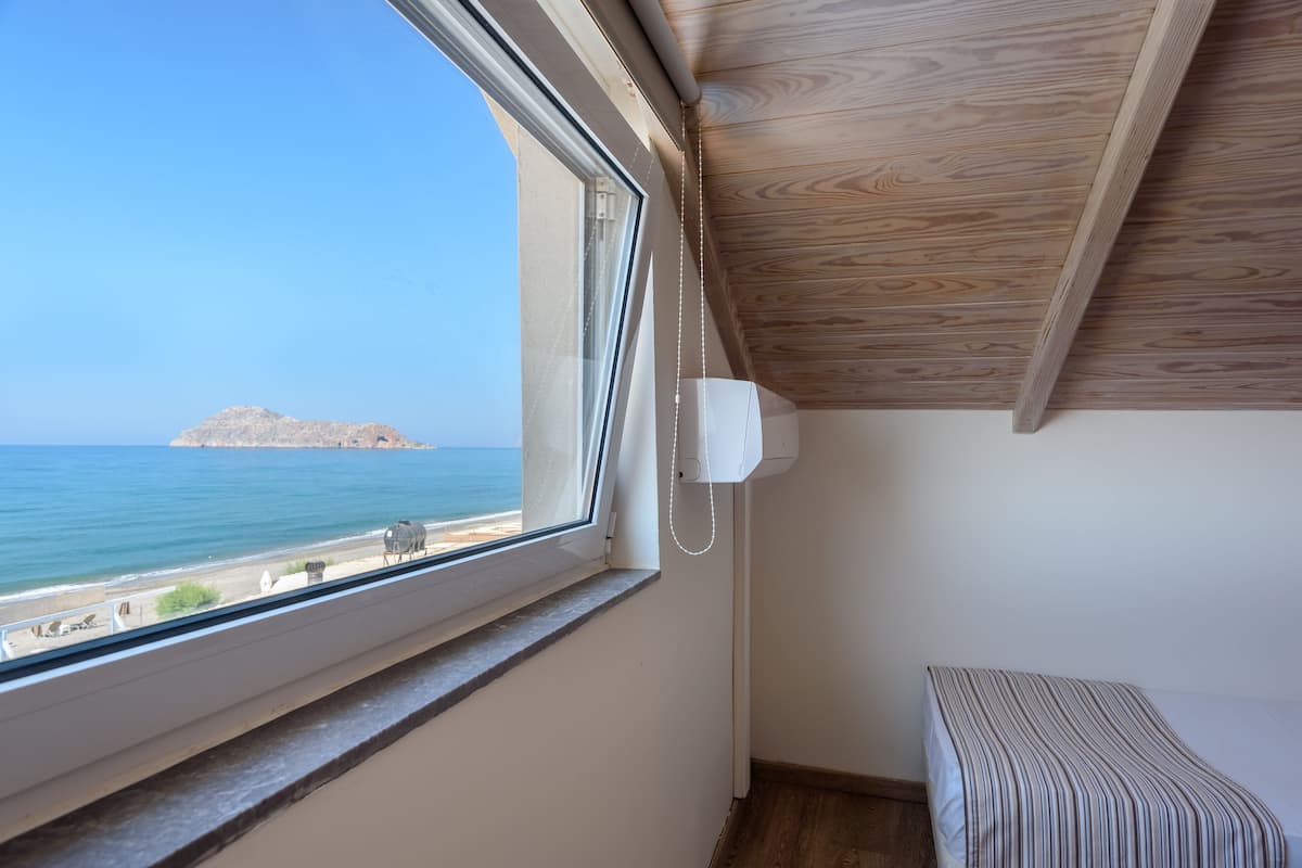 A room with a window overlooking a beach, a bed, and a ceiling with wooden paneling.