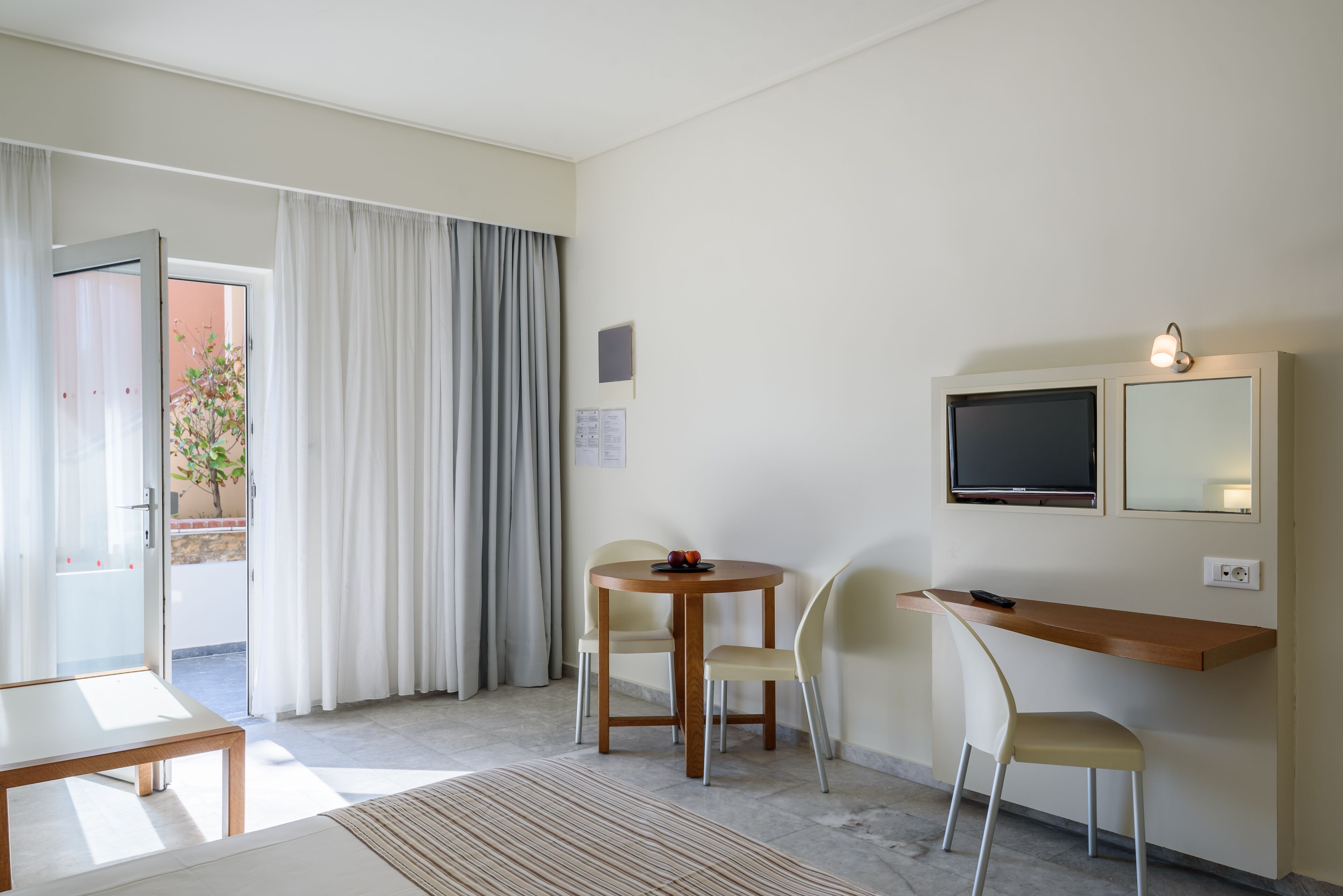 A hotel room with a dining area, a TV mounted on the wall, and a sliding glass door leading to an outdoor area.