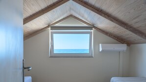 A room with a sloped wooden ceiling, a window with a view of the sea, a bed, and an air conditioning unit.