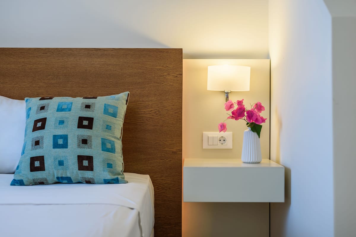 A neatly made bed with a patterned pillow, a bedside table with a lamp and a vase of flowers, and a wall socket.