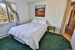 A bedroom with a bed, a green bedside table, a painting on the wall, and a window with curtains. - Cozy Cabin bordering Pikes National Forest  (Woodland Park)