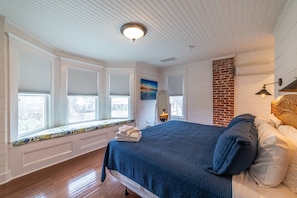 A bedroom with a large bed, a brick accent wall, a window seat with a view, and a ceiling light fixture.