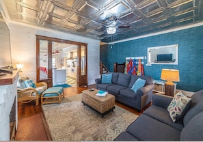 A living room with a dark gray sofa, wicker ottoman, and a blue rug. There are flags on the wall and a chandelier.