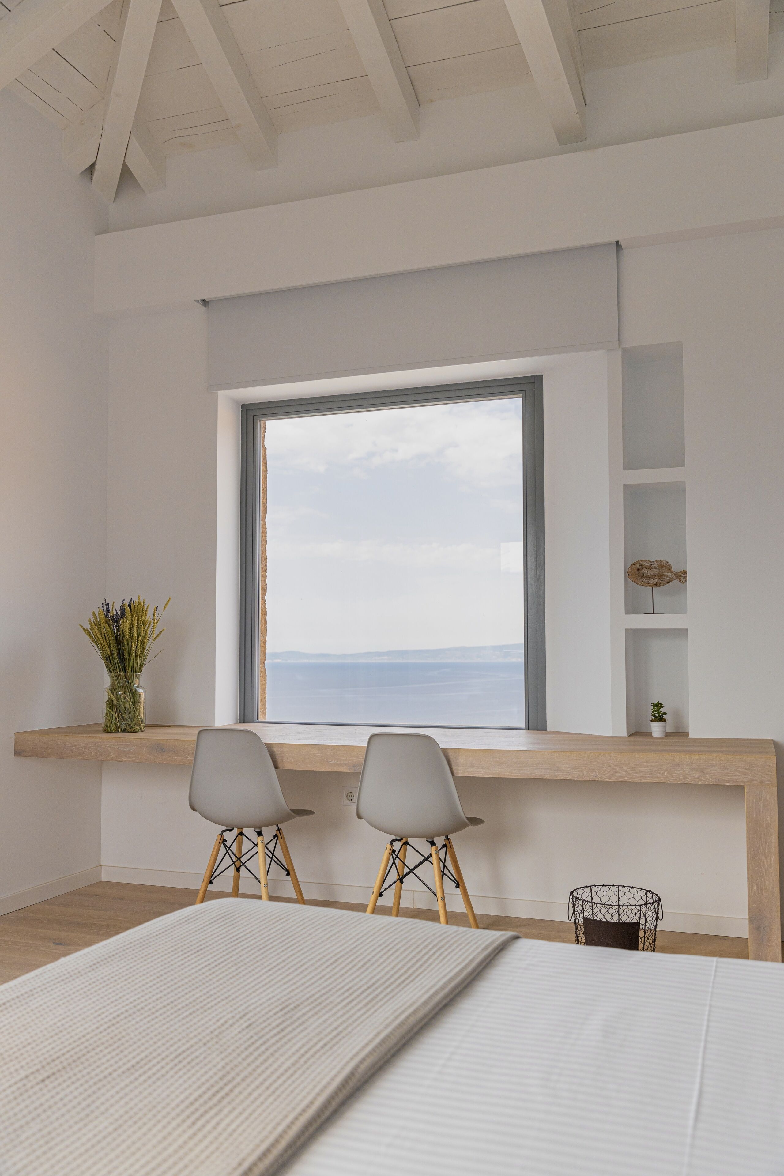 Una habitación minimalista con un ventanal grande, dos sillas, una mesita y una cama.
