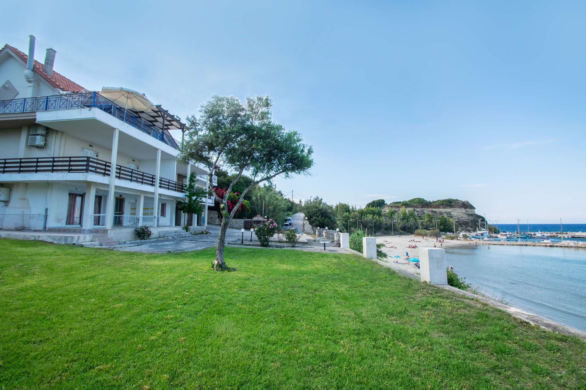A coastal property with a large building featuring multiple balconies, a grassy lawn, and a beach with boats.