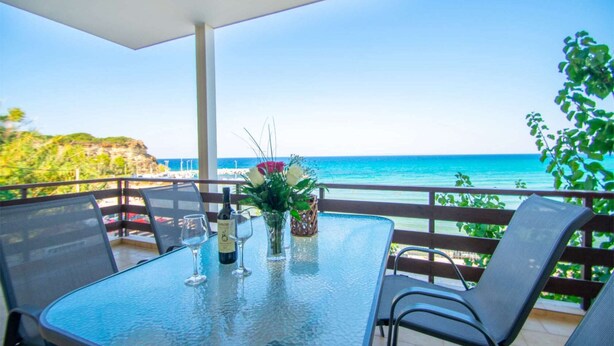 A table with a glass top, two chairs, a bottle of wine, and a vase with flowers on a balcony overlooking the sea.