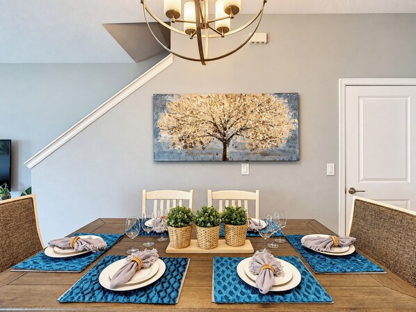A dining area with a table set for four, a chandelier, and a painting of a tree on the wall.