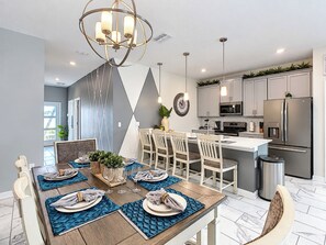 A modern kitchen with a dining area set for six, featuring a central island, stainless steel appliances, and a chandelier.