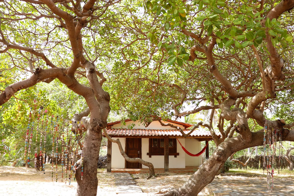 Uma casa com telhado de cerâmica e porta de madeira, cercada por árvores e uma rede.