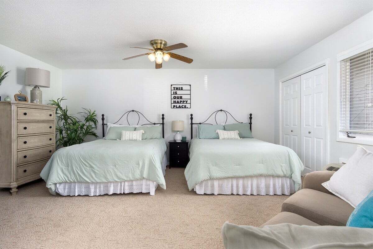 Une chambre avec deux lits, un ventilateur de plafond, une commode, une table de chevet, une lampe et une fenêtre avec des stores.