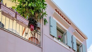 Un immeuble de deux étages avec une façade rose, de nombreuses fenêtres dotées de volets et un balcon orné de plantes en pot.