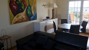 Una sala de estar con un sofá negro, una mesa de comedor con sillas, un cuadro de jarrones de colores y una ventana con vista a edificios.