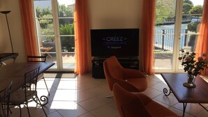 Una sala de estar con televisor, cortinas naranjas, una mesa de comedor y vistas a un jardín y un cuerpo de agua.