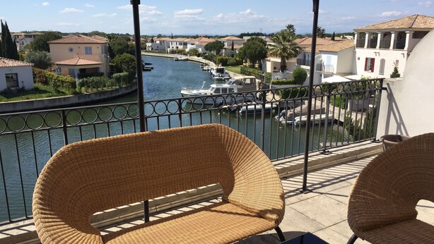 Un balcón con sillas de mimbre y una mesa con vistas a un canal con barcos y casas.