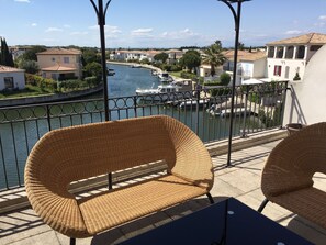 A balcony with wicker chairs and a table overlooking a canal with boats and houses.