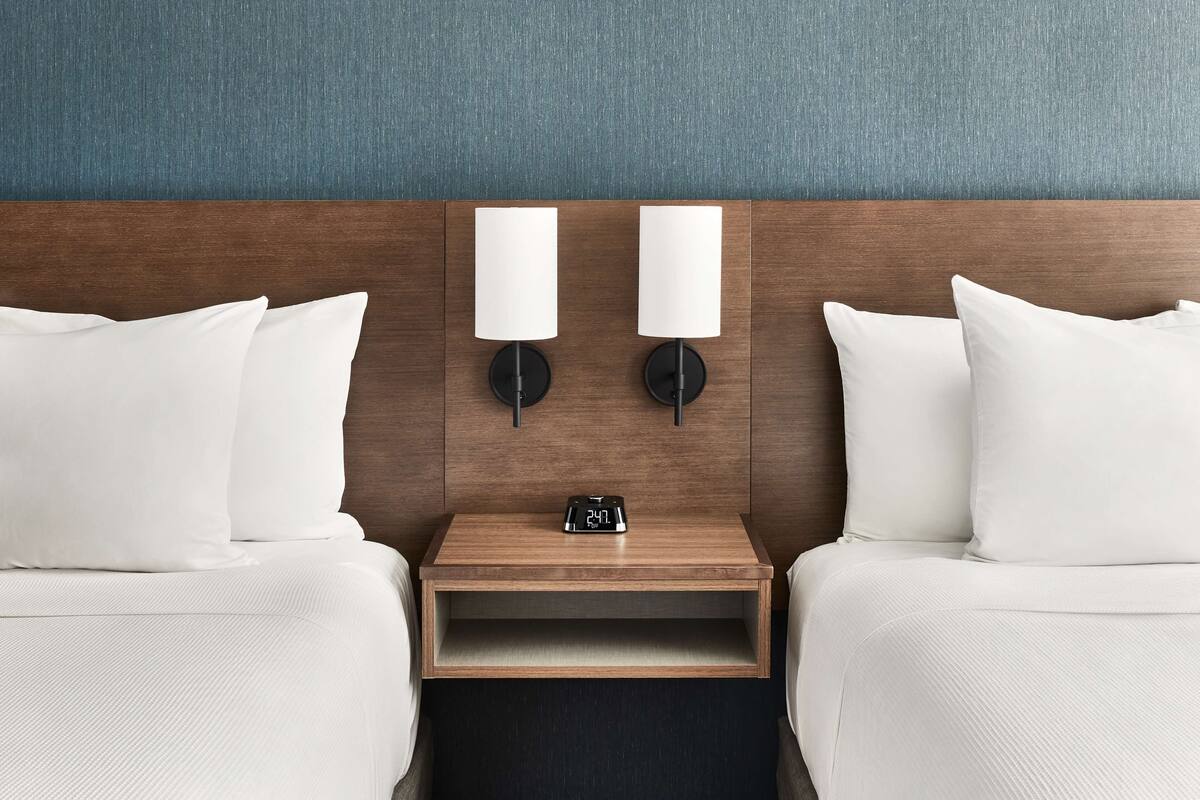 A hotel room with two beds, a nightstand with a clock, and wall-mounted lamps.