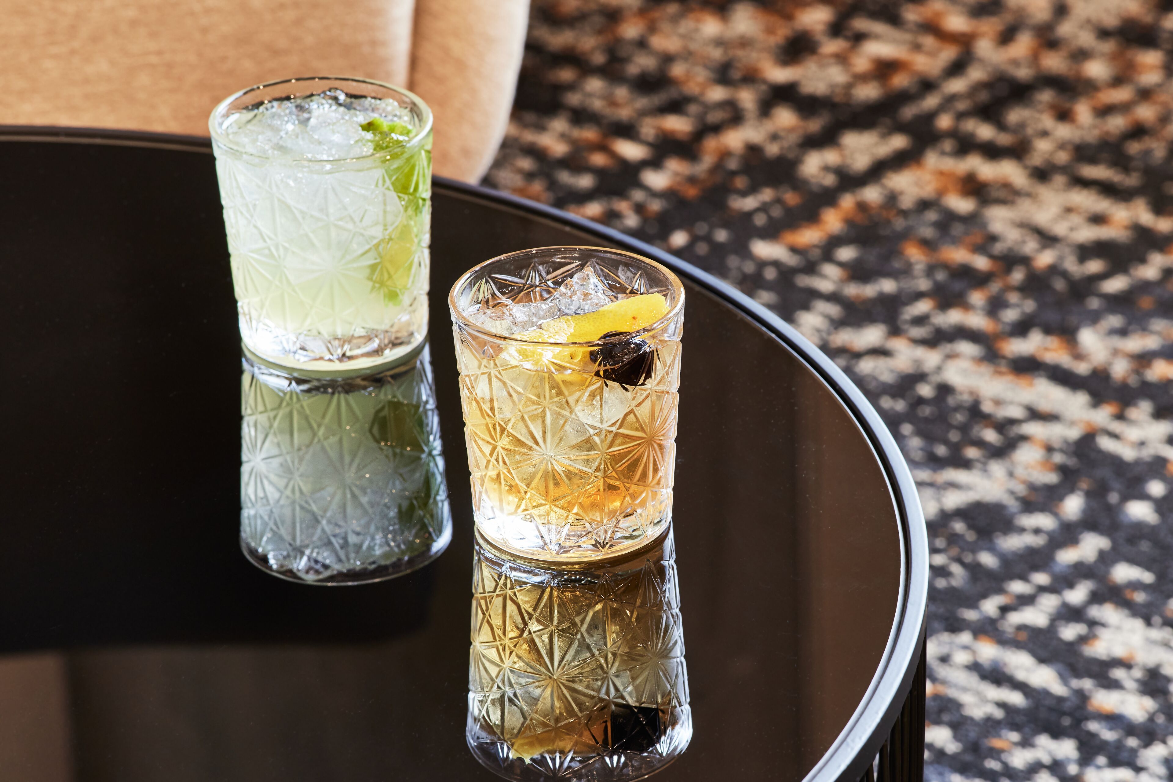 Two glasses of cocktails on a reflective black table with a textured surface.