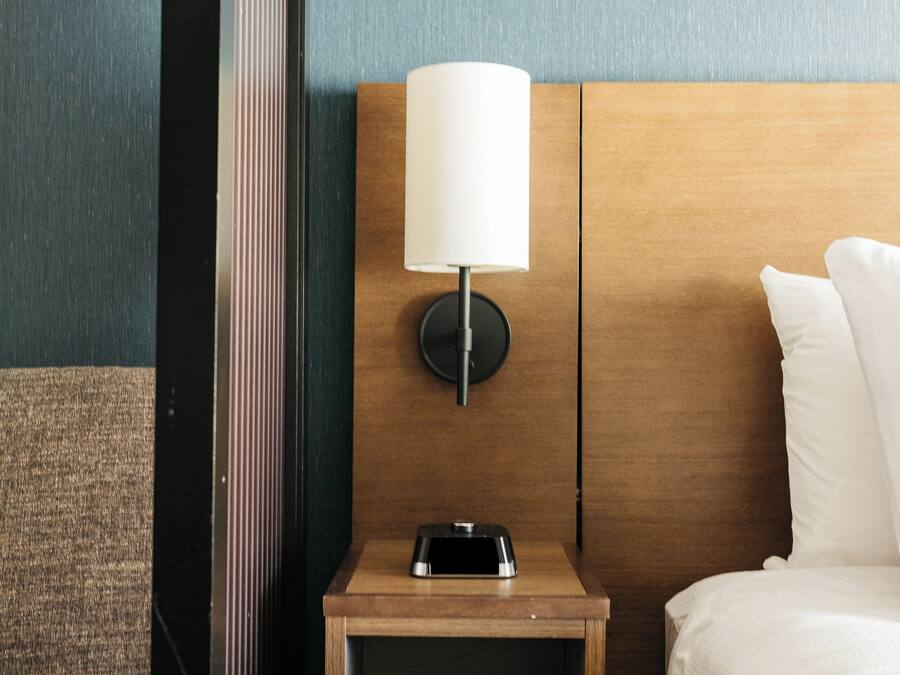 Una habitación de hotel ordenada con una mesita de noche de madera, una lámpara y una cama con ropa blanca.