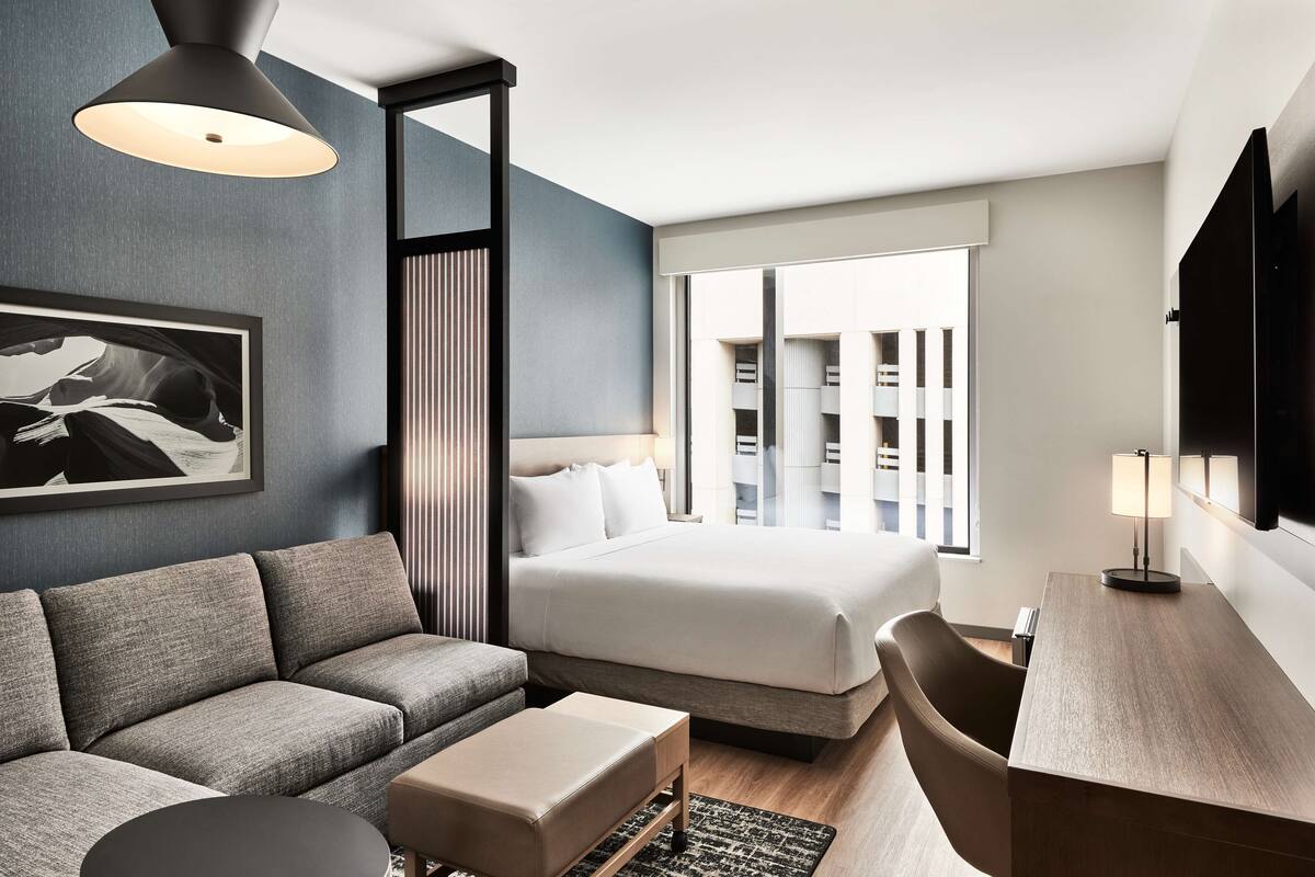 A modern hotel room with a sofa, a bed, a desk, and a large window.