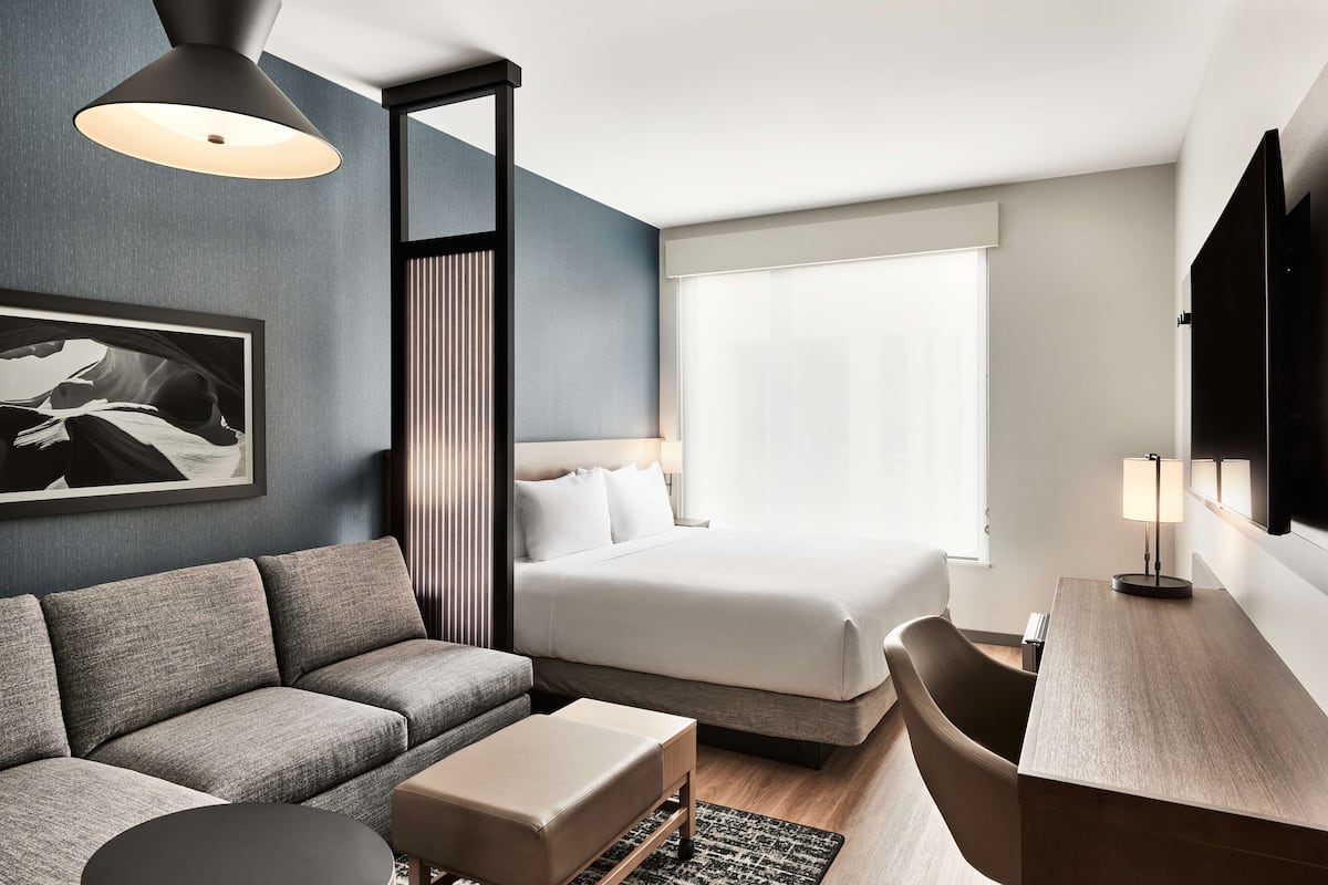 A modern hotel room with a bed, sofa, desk, and a large window.