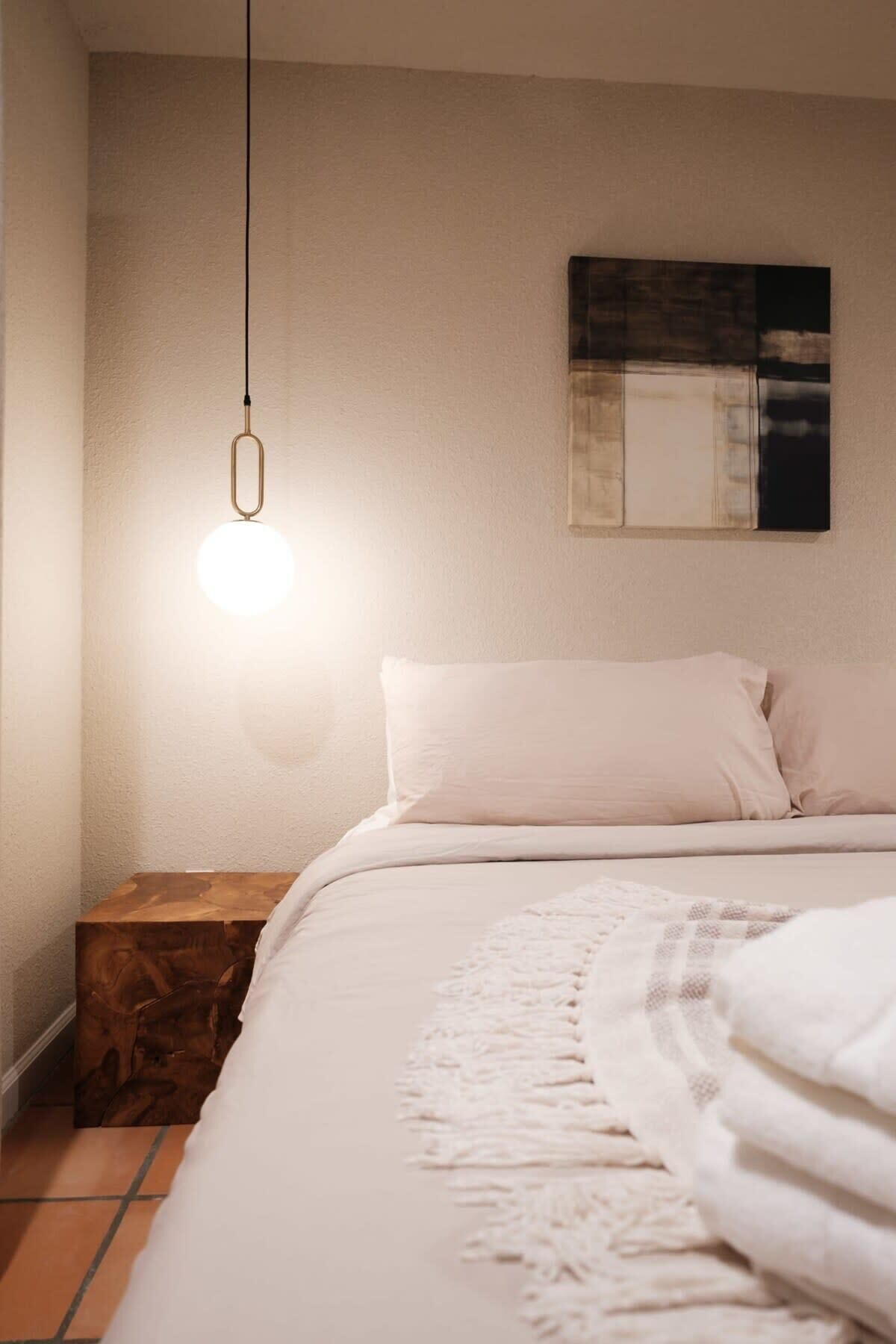 A bedroom with a bed, a bedside table, a hanging light fixture, and a wall-mounted artwork.