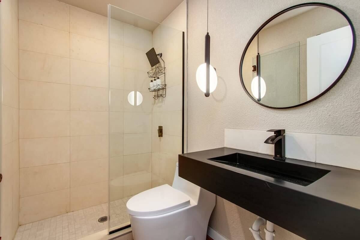 Un baño moderno con cabina de ducha de vidrio, inodoro blanco, lavabo negro y espejo redondo.