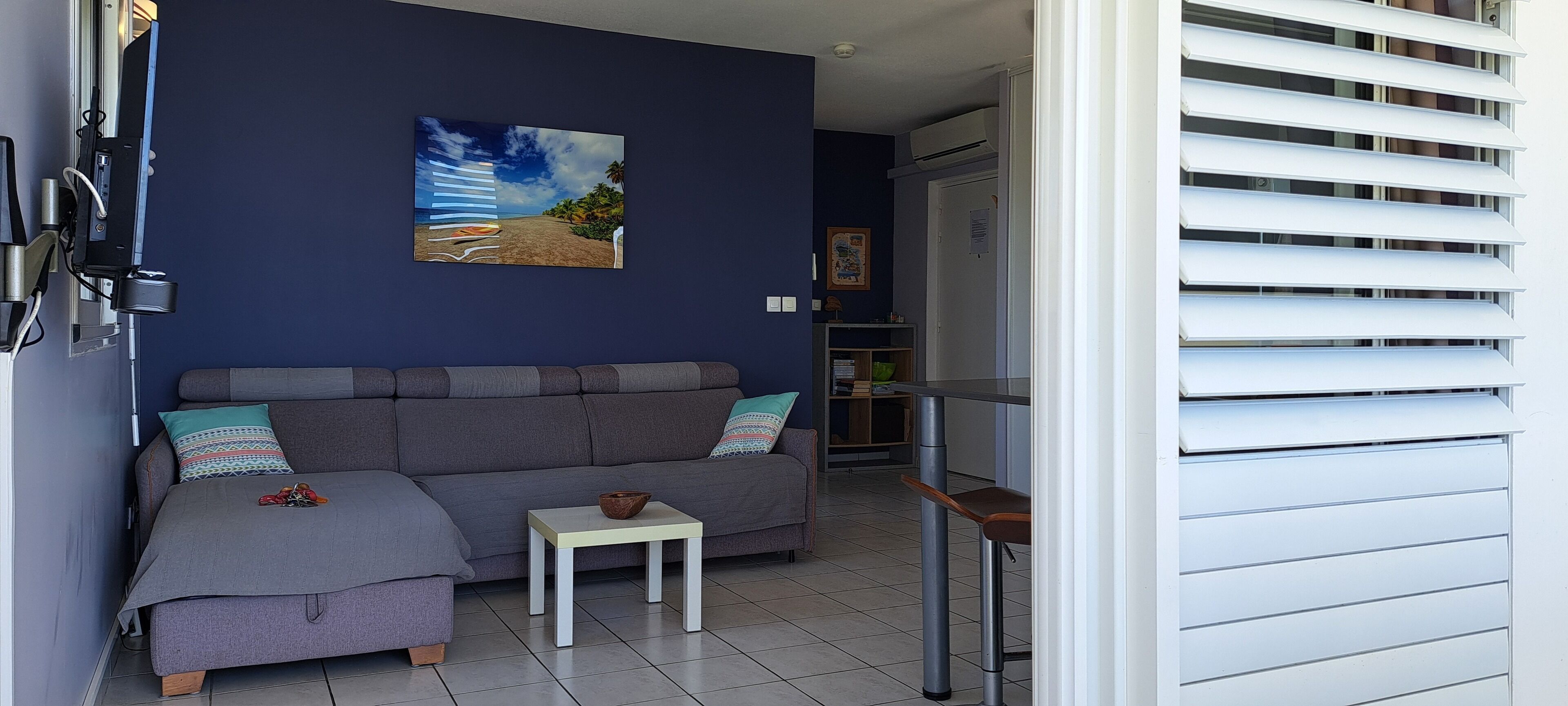 Uma sala de estar com um sofá cinza, uma mesinha e um quadro de uma paisagem praiana.