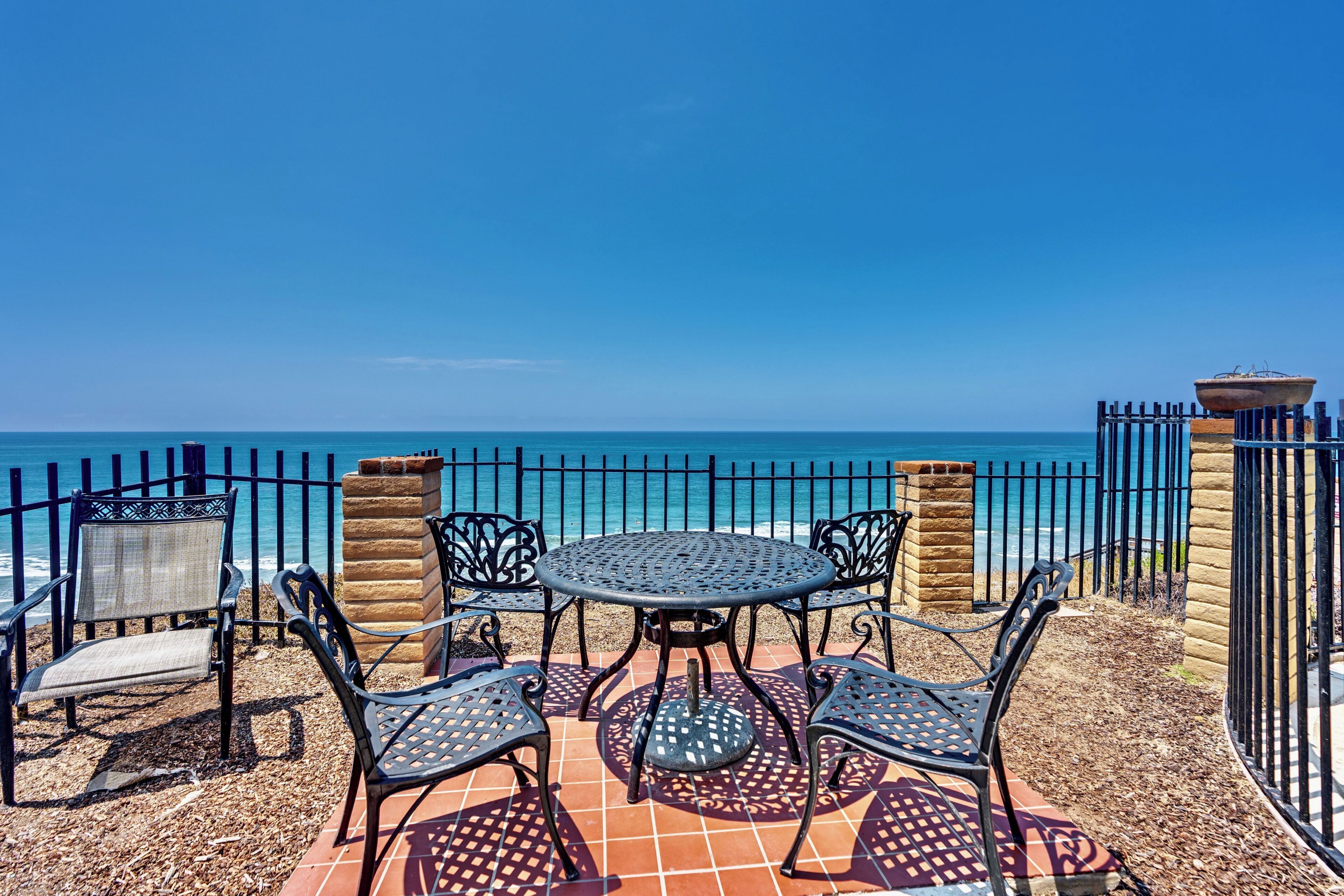 Ein Außenbereich mit Tisch und Stühlen, Blick auf das Meer und einem Metallzaun.