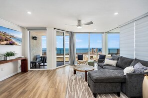 Ein großzügiges Wohnzimmer mit grauem Sofa, einem Couchtisch und einem Fernsehgerät an der Wand. Ein Balkon bietet Blick aufs Meer.