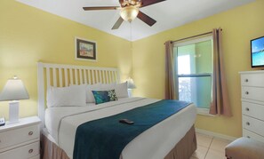 Habitación de hotel con cama, mesitas de noche, ventilador de techo, ventana con cortinas y televisor en la pared.