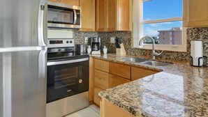 A modern kitchen with stainless steel appliances, granite countertops, wooden cabinets, and a window with a city view. - Treasure Island Beachfront-Top Floor End Unit  Stunning Sunsets South Beach 601 (Treasure Island)