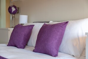 Una cama con almohadas blancas y púrpuras, un globo terráqueo y una lámpara sobre una mesita de noche.